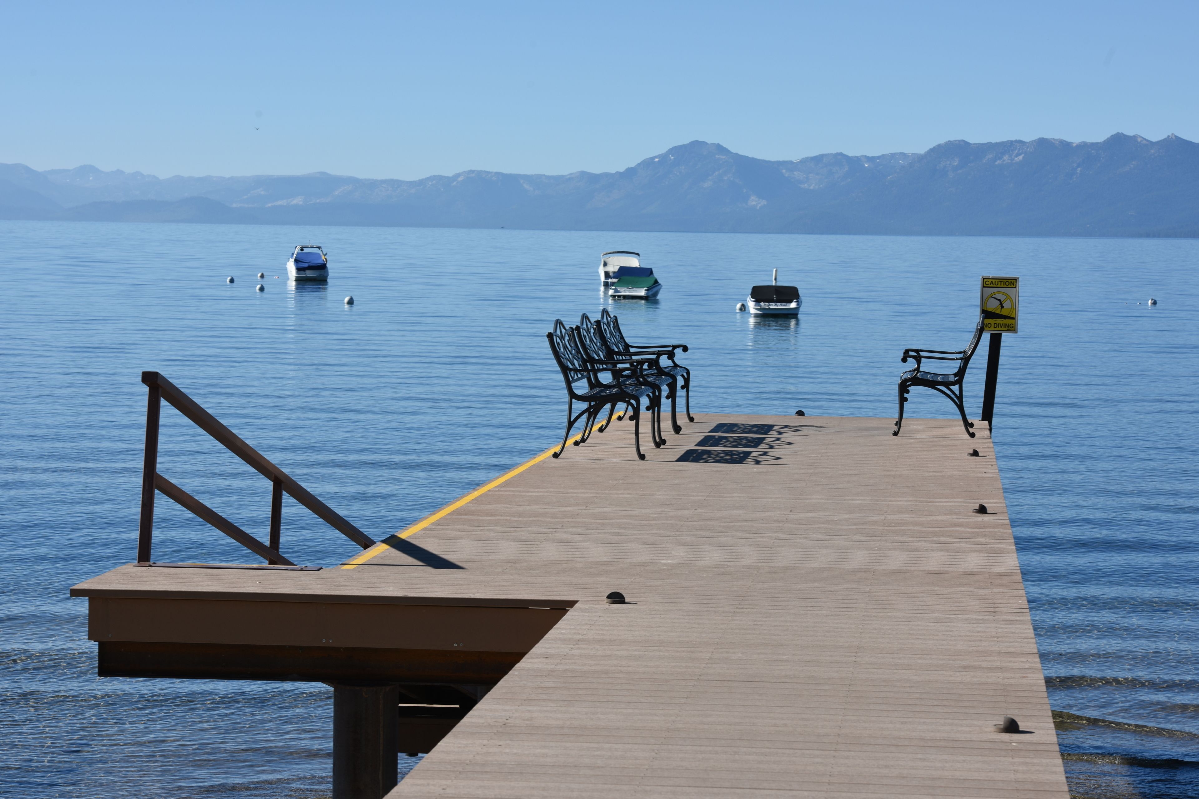 Un muelle de madera con bancos de metal y un cartel de advertencia, que se adentra en un lago tranquilo con barcos y montañas al fondo.