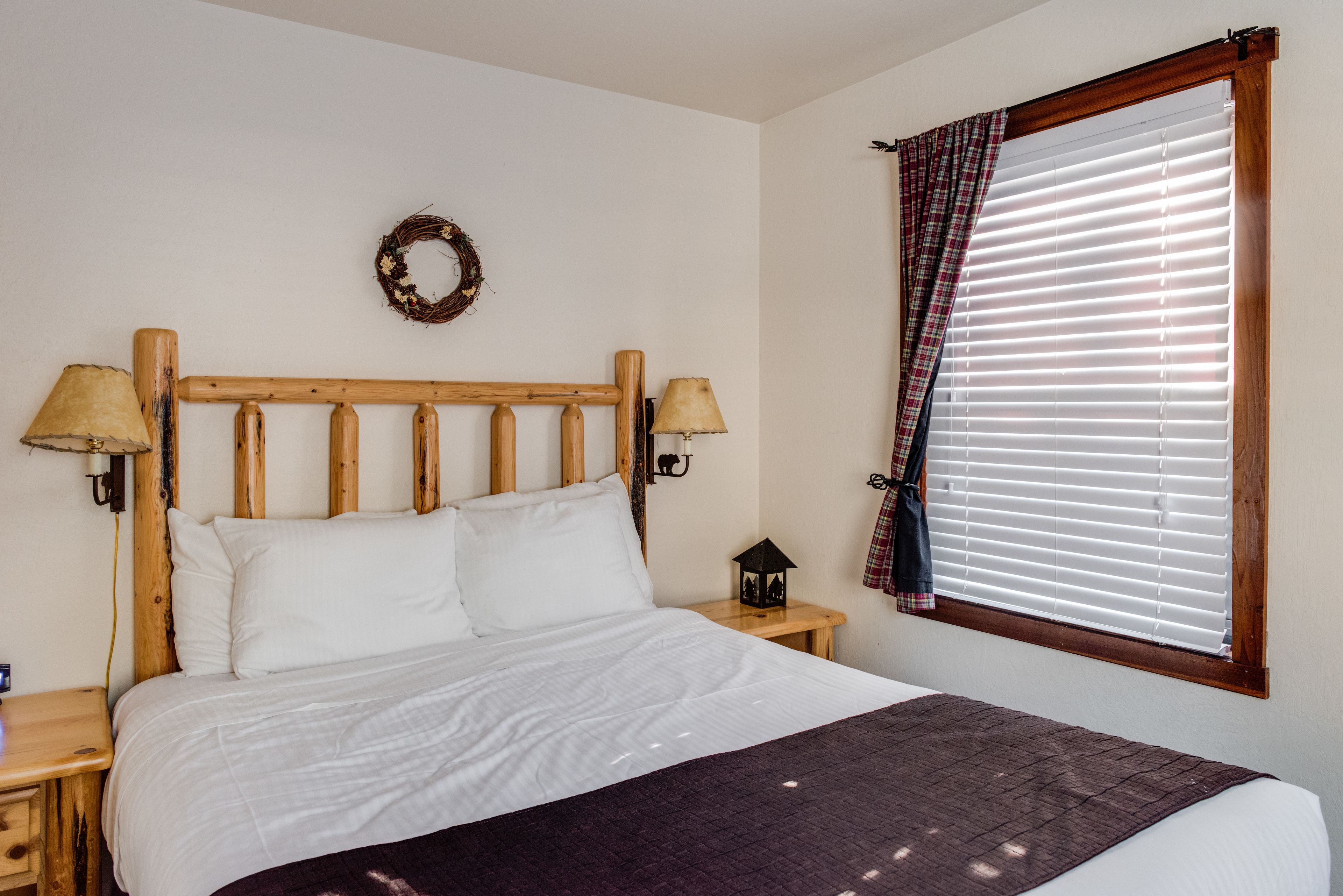 Quarto com cabeceira de madeira, duas luminárias de cabeceira, mesa de cabeceira com um candeeiro, janela com persianas e uma coroa de flores pendurada na parede.