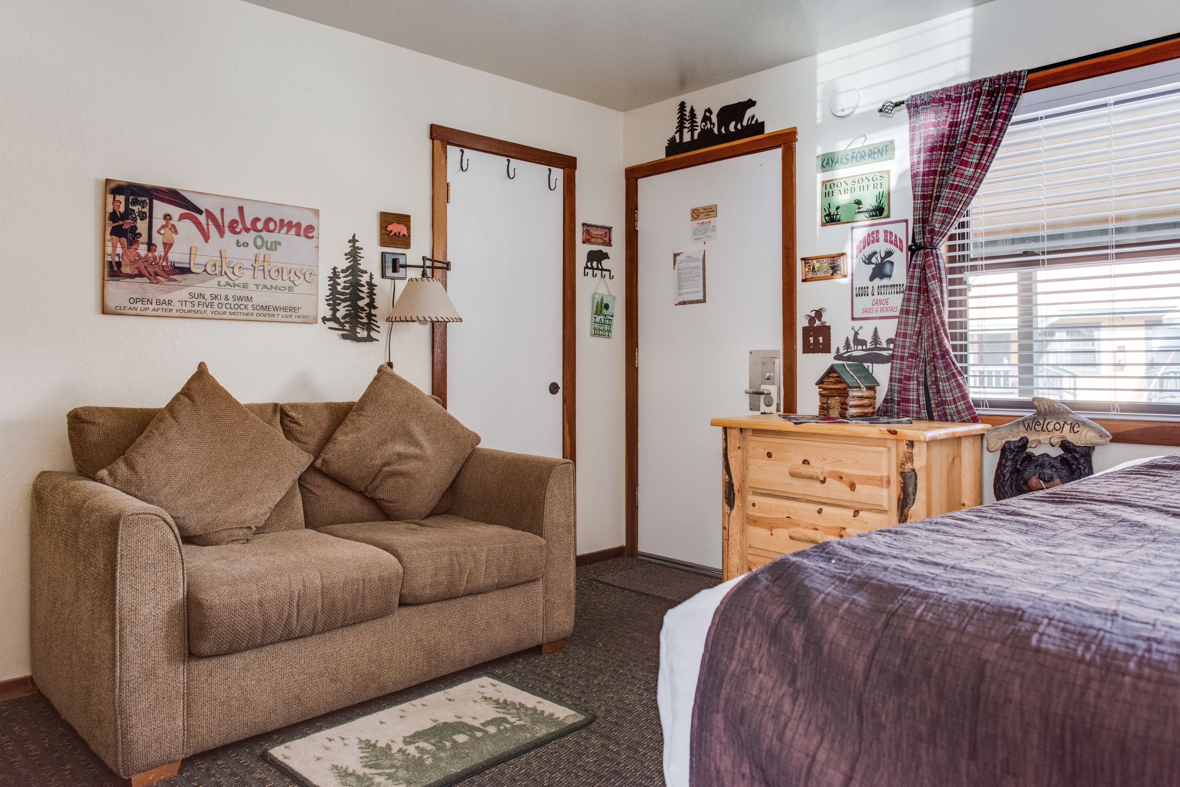 A room with a brown sofa, a wooden dresser, a bed with a purple bedspread, and a window with blinds.
