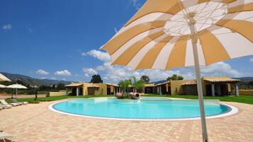 A swimming pool with a tiled deck, surrounded by lounge chairs and umbrellas, with buildings in the background.