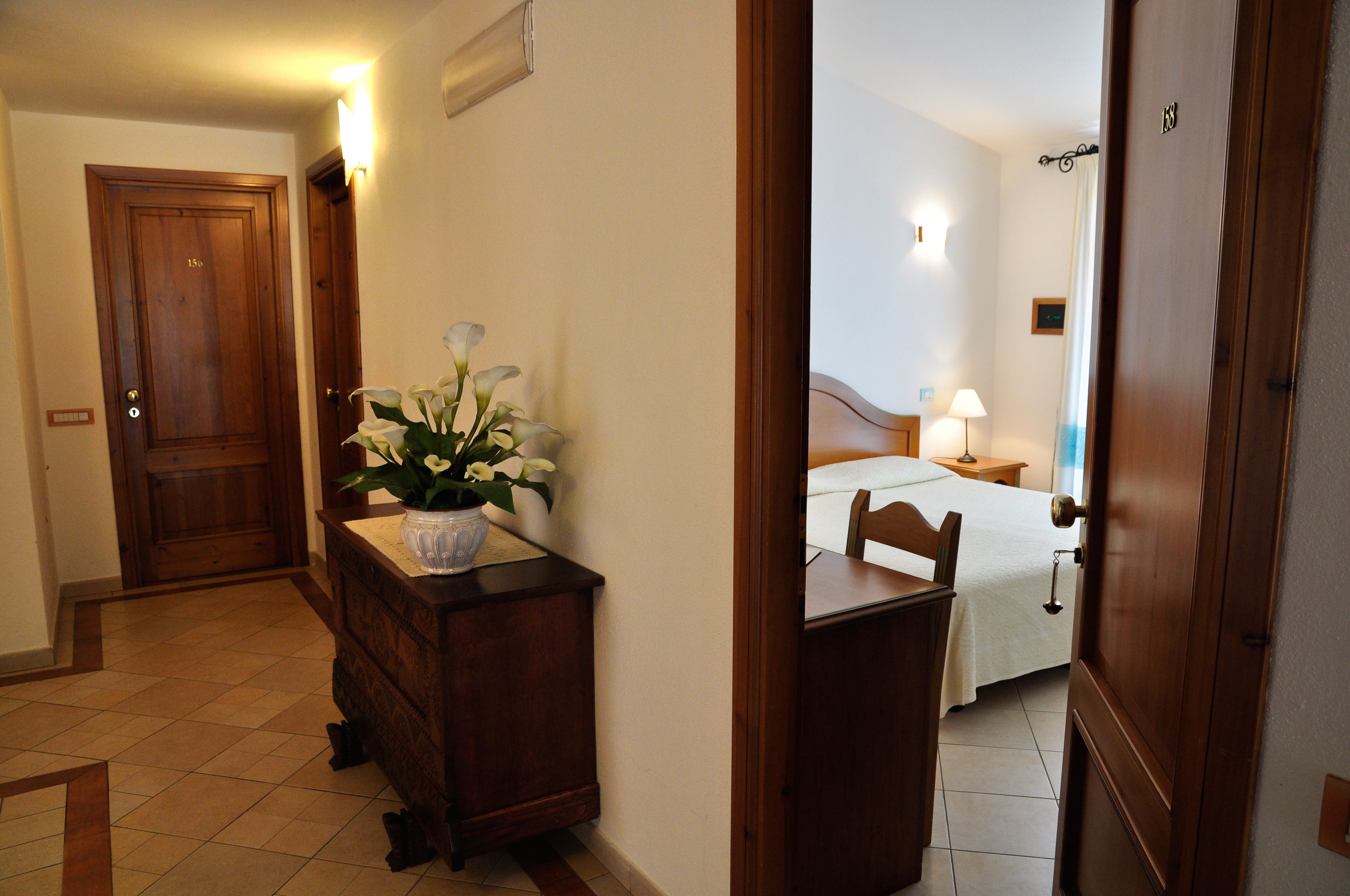 Quarto de hotel com cômoda de madeira, uma cama com colcha branca, mesa de cabeceira e abajur, e porta de madeira.