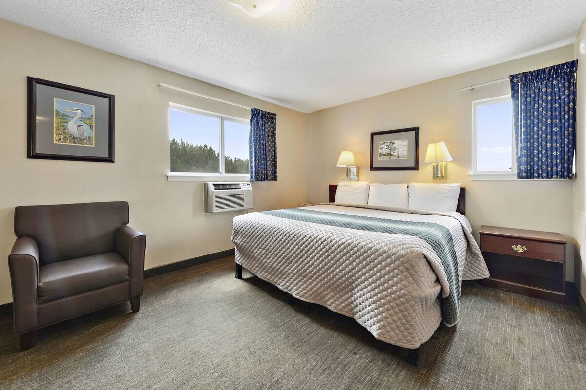 A hotel room with a bed, a chair, a nightstand, and two windows with curtains.