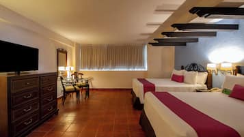 Habitación de hotel con televisor, escritorio y silla, dos camas con almohadas y ventana con cortinas.