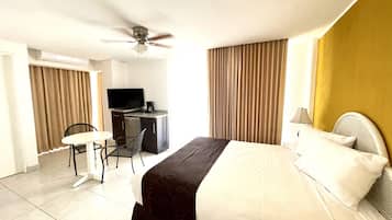 Habitación de hotel con cama, escritorio con televisor, silla y ventilador de techo.