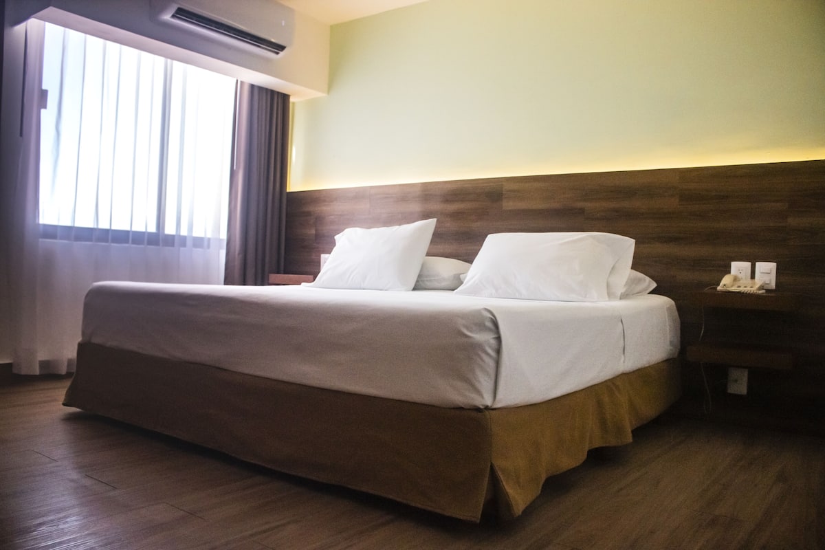 Une chambre d’hôtel avec un grand lit, une tête de lit en bois, une table de chevet avec une lampe et une fenêtre avec des rideaux.