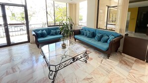 A spacious living area with a blue sofa, a glass coffee table, and large windows offering a view of the outdoors. - Hotel del Paseo Campeche (Campeche)