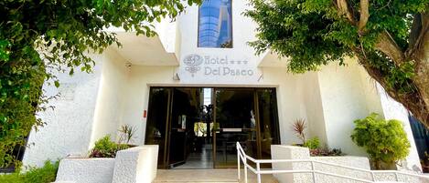 A entrada do Hotel Del Paseo, com uma escadaria que leva à entrada, rodeada por vegetação.