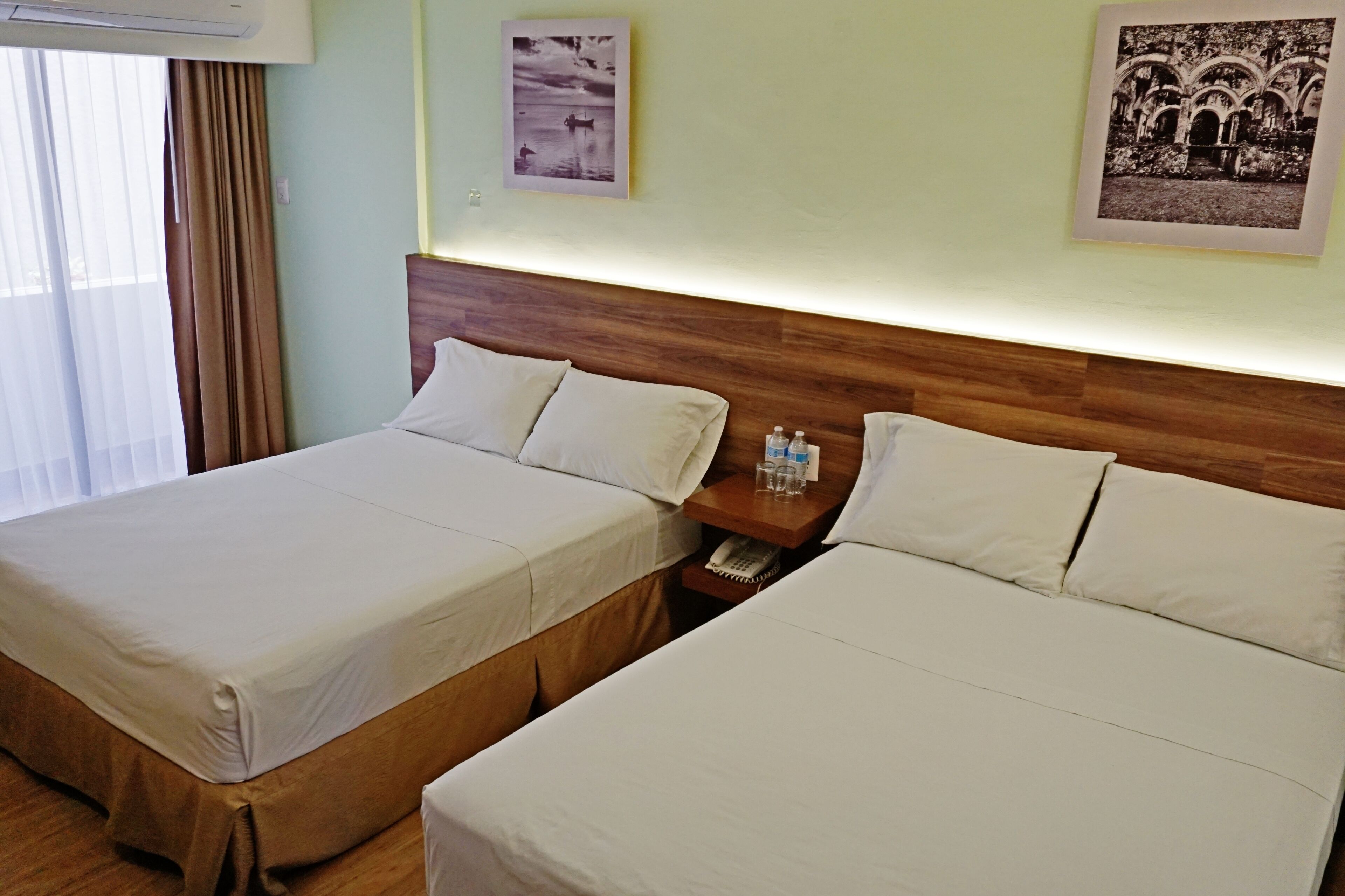 A hotel room with two beds, a wooden headboard, a nightstand with a phone and water bottles, and two framed pictures on the wall.