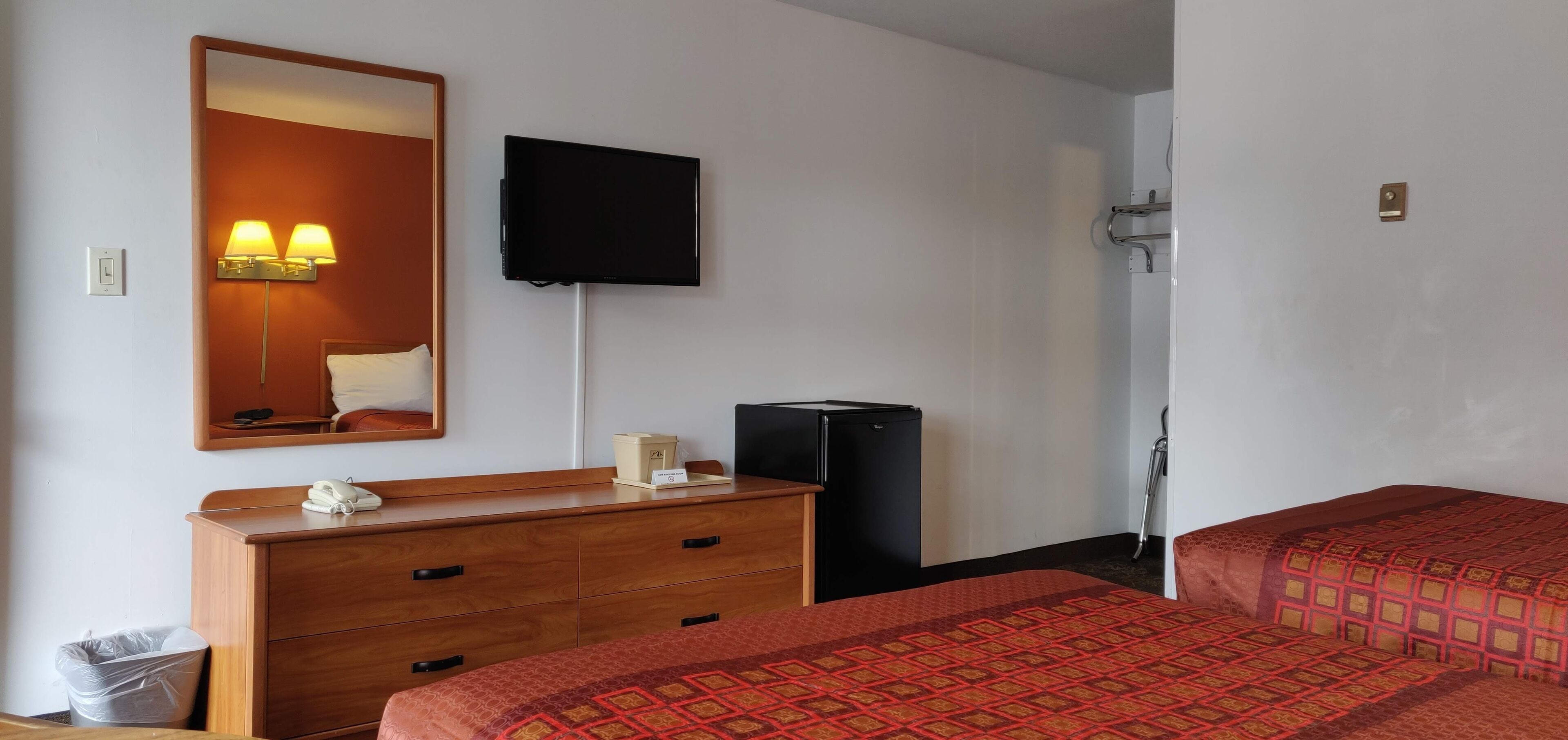 A hotel room with a wooden dresser, a flat-screen TV mounted on the wall, a bed with a patterned bedspread, and a small trash bin.