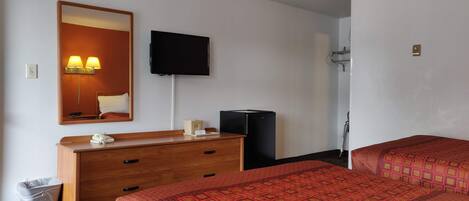A hotel room with a wooden dresser, a flat-screen TV mounted on the wall, a bed with a patterned bedspread, and a small trash bin.