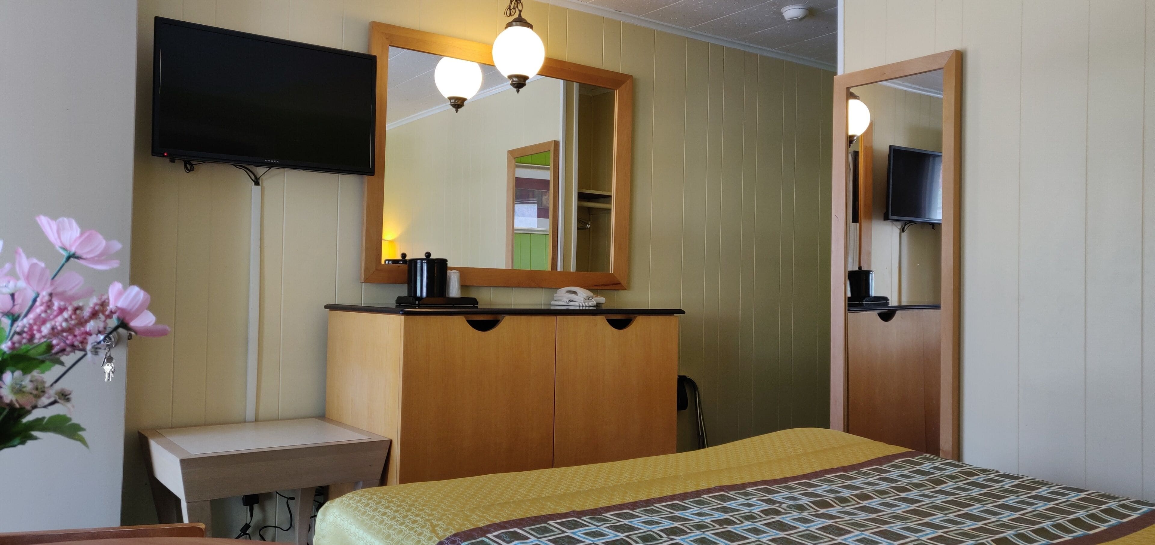Une chambre d’hôtel comprenant un lit, une armoire en bois, une table de chevet, un miroir, un téléviseur fixé au mur et un arrangement floral.