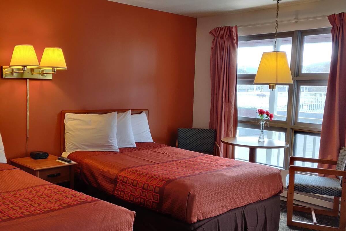 Quarto de hotel com duas camas, mesa de cabeceira com abajur, uma cadeira e janela com cortinas.