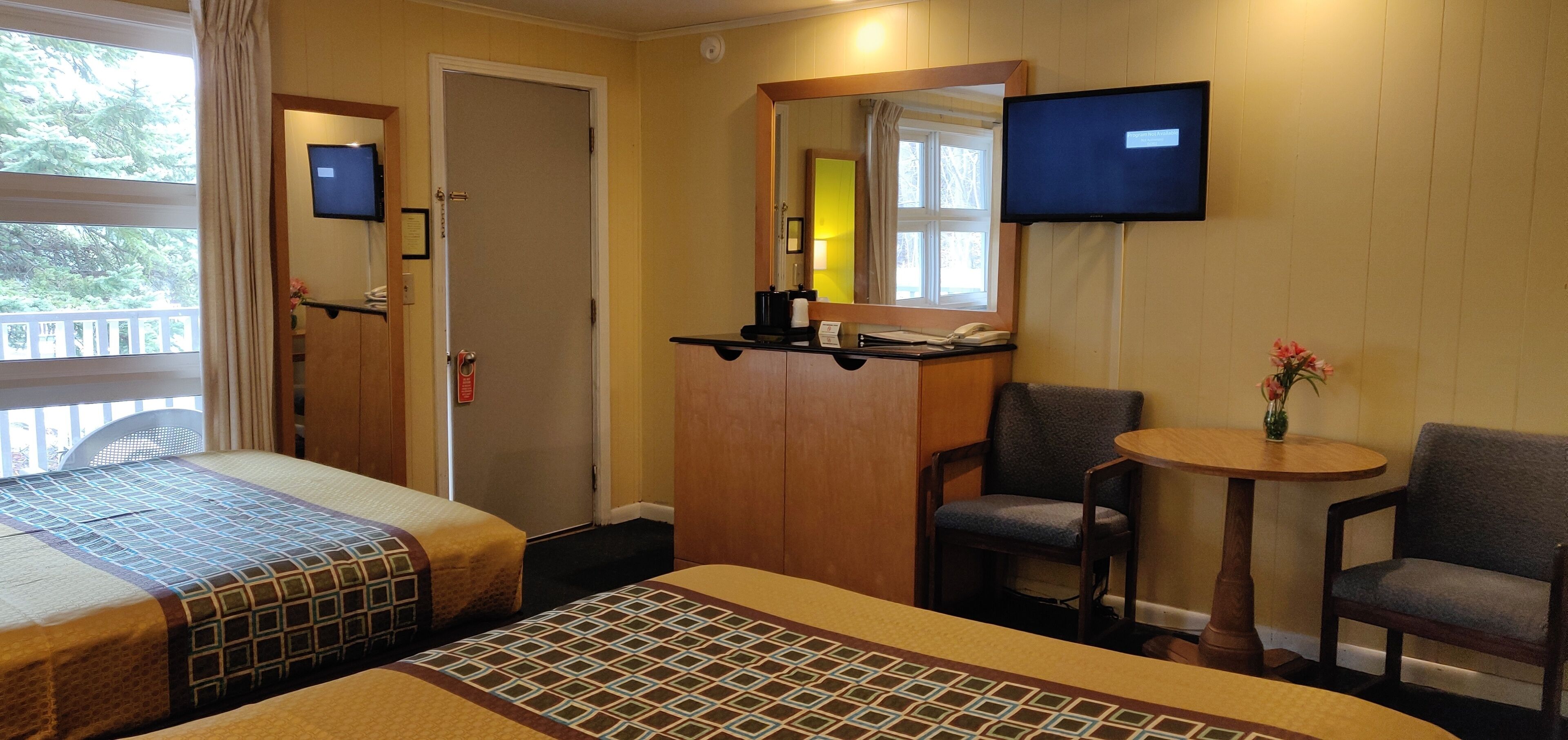A hotel room with two beds, a small table with a chair, a TV mounted on the wall, and a window with a view of trees.