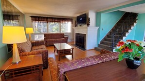 Sala de estar con sofá, mesa de centro y chimenea. Se ve una escalera a la derecha y un televisor sobre la chimenea.