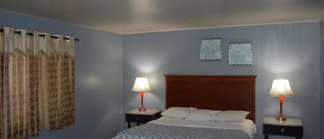 A hotel room with a bed, two bedside tables with lamps, a window with curtains, and a wall-mounted air conditioning unit.