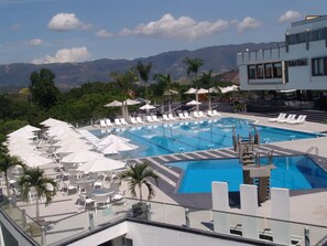 A pool area with lounge chairs and umbrellas, surrounded by palm trees and mountains in the background.