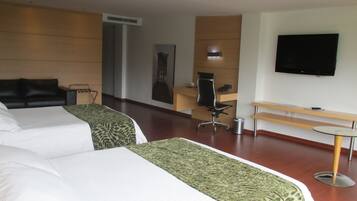 A hotel room with two beds, a desk, a chair, a trash bin, a bench, and a TV mounted on the wall.