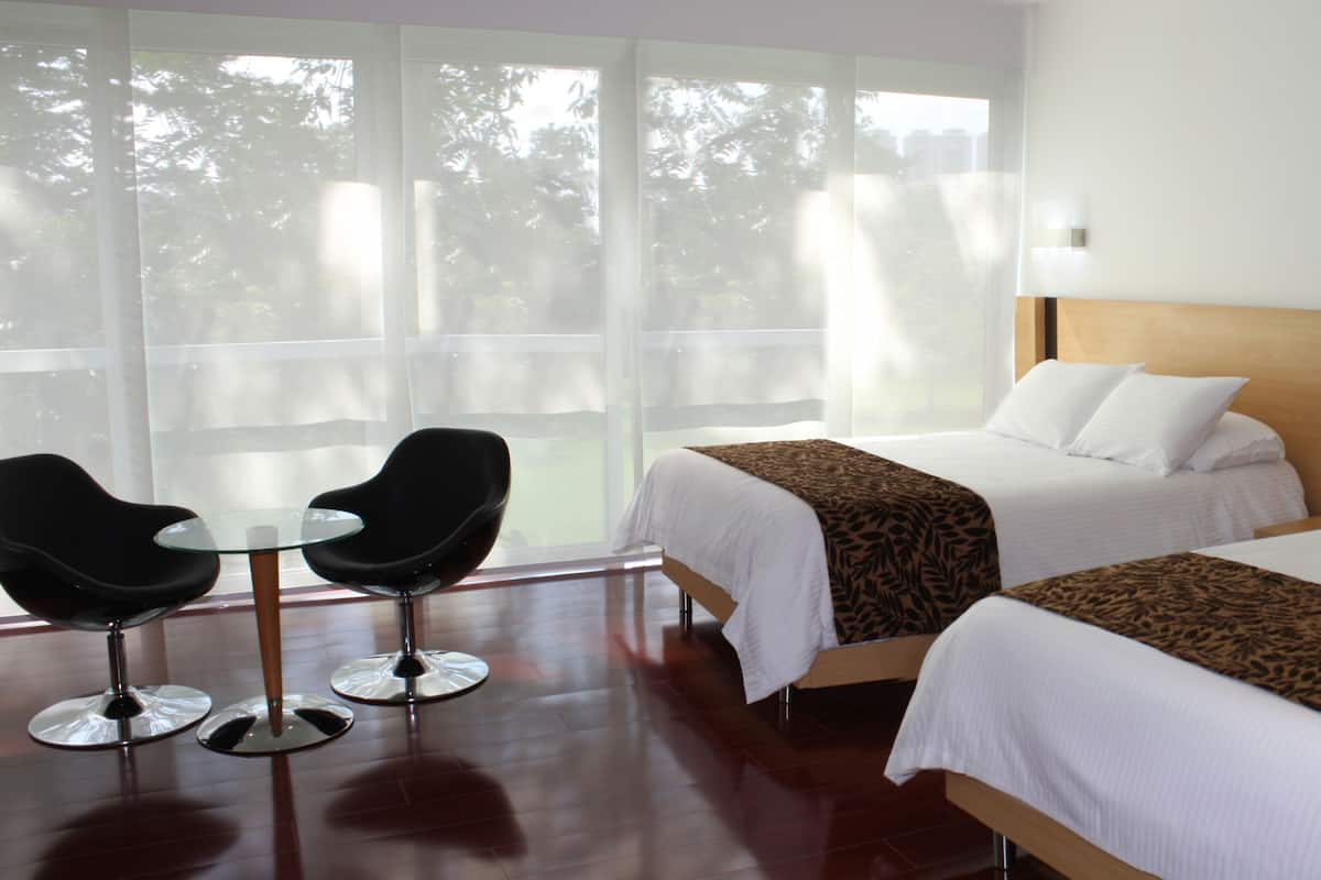 Una habitación de hotel con dos camas, una mesa de vidrio con sillas y ventanas grandes con cortinas traslúcidas.