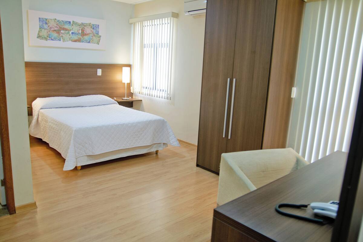 A hotel room with a bed, bedside table, lamp, wardrobe, and a desk with a telephone.
