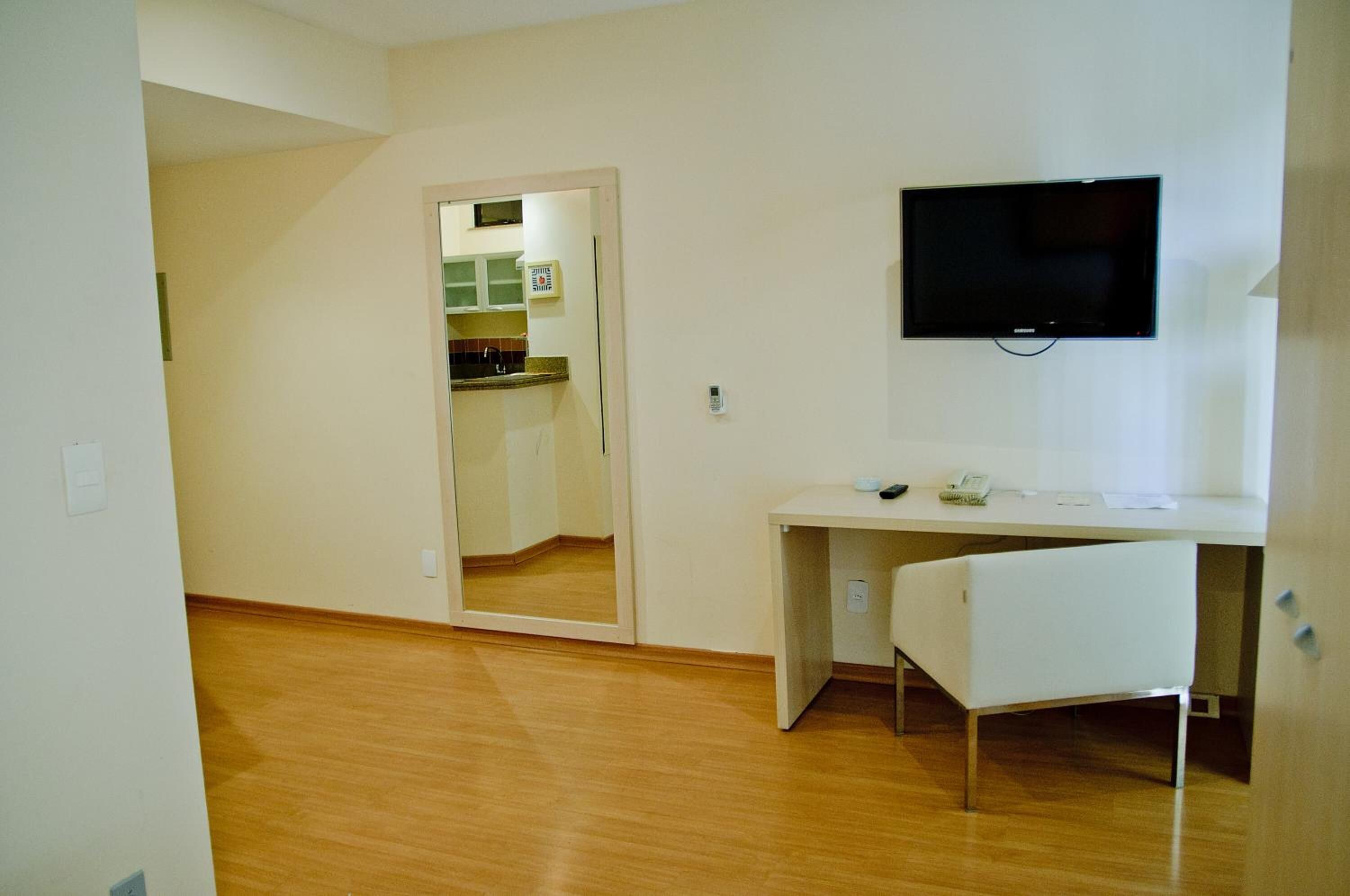A room with a wooden floor, a white desk with a chair, a flat-screen TV mounted on the wall, and a door leading to a kitchen area.