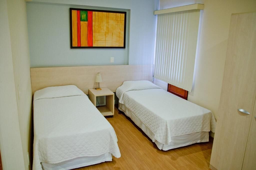 A small, clean room with two single beds, a nightstand with a lamp, and a window with blinds.