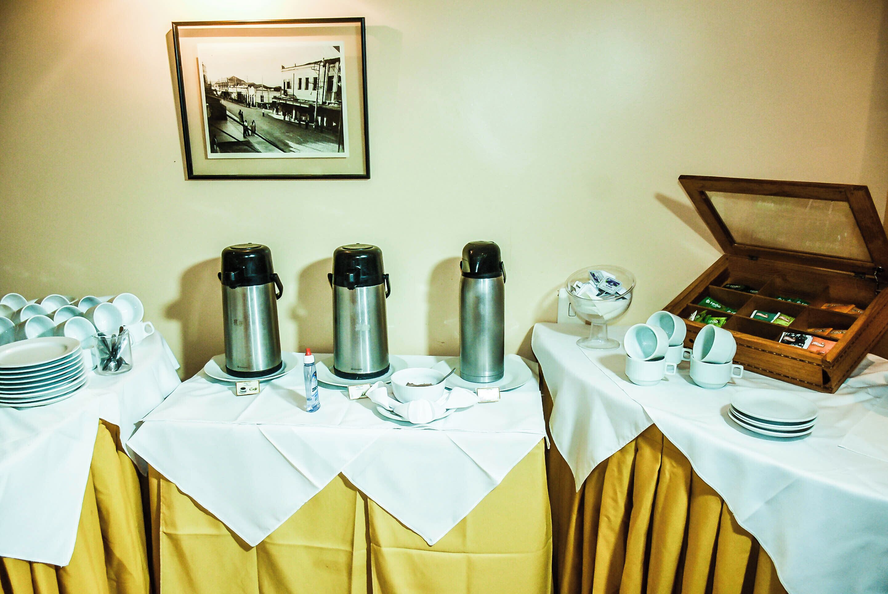 A breakfast buffet setup with coffee makers, plates, cups, and a box of tea.