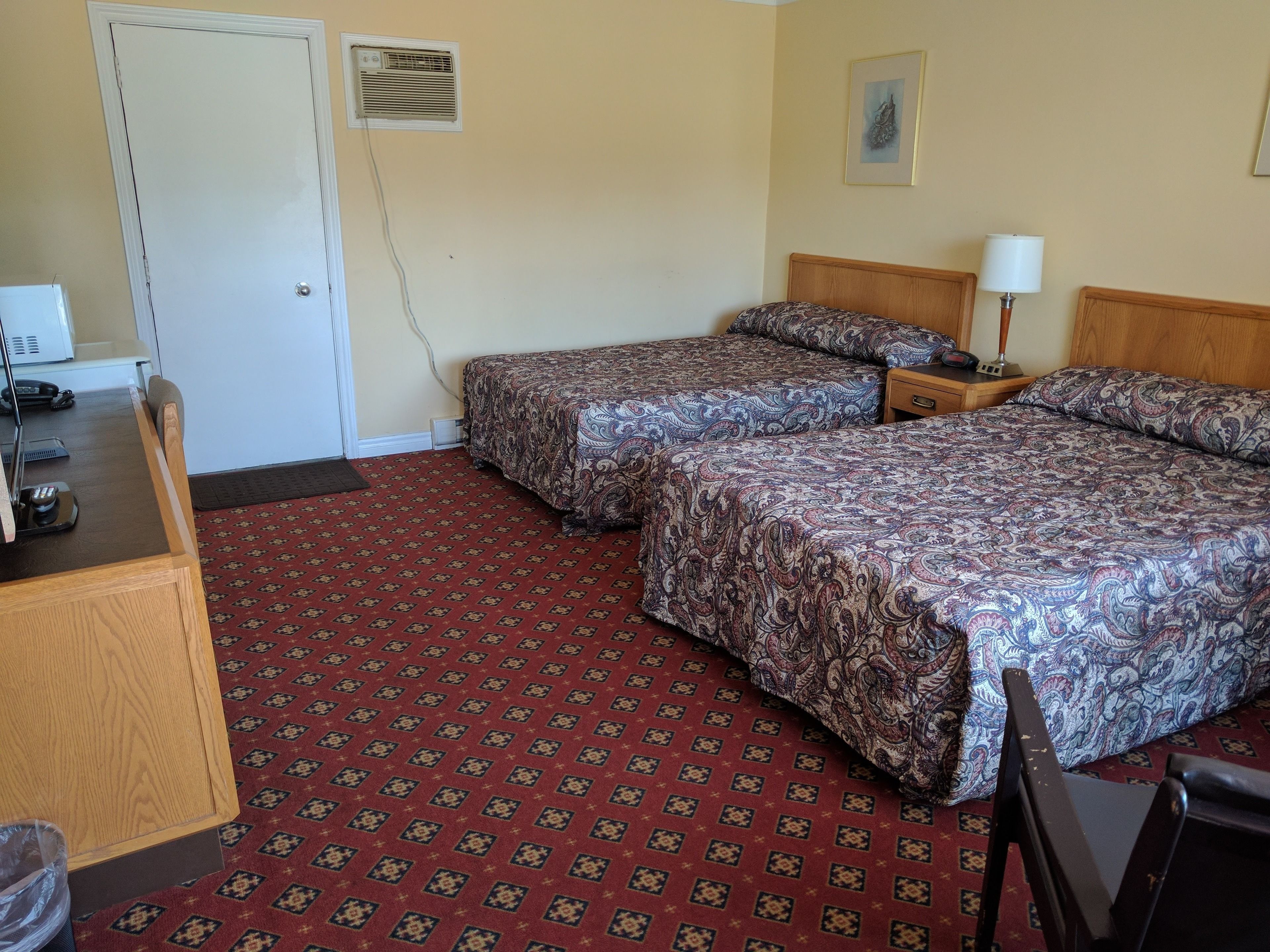A hotel room with two beds, a desk, a lamp, and a door.