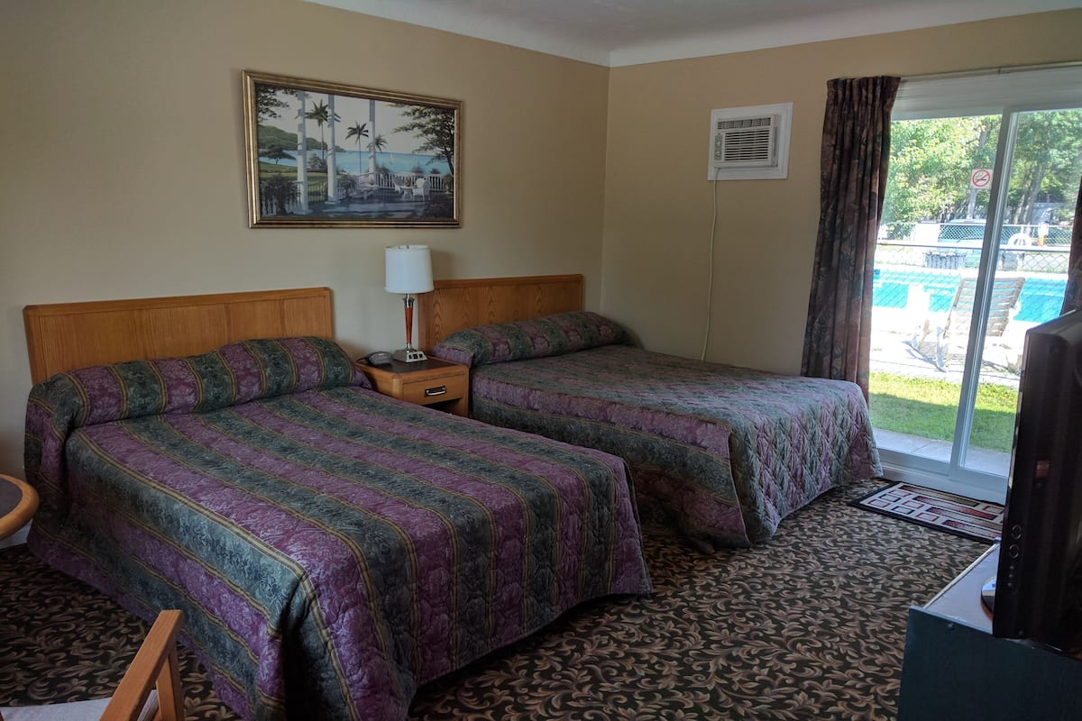 A hotel room with two beds, a wooden headboard, a nightstand with a lamp, a painting on the wall, and a view of a pool through the window.