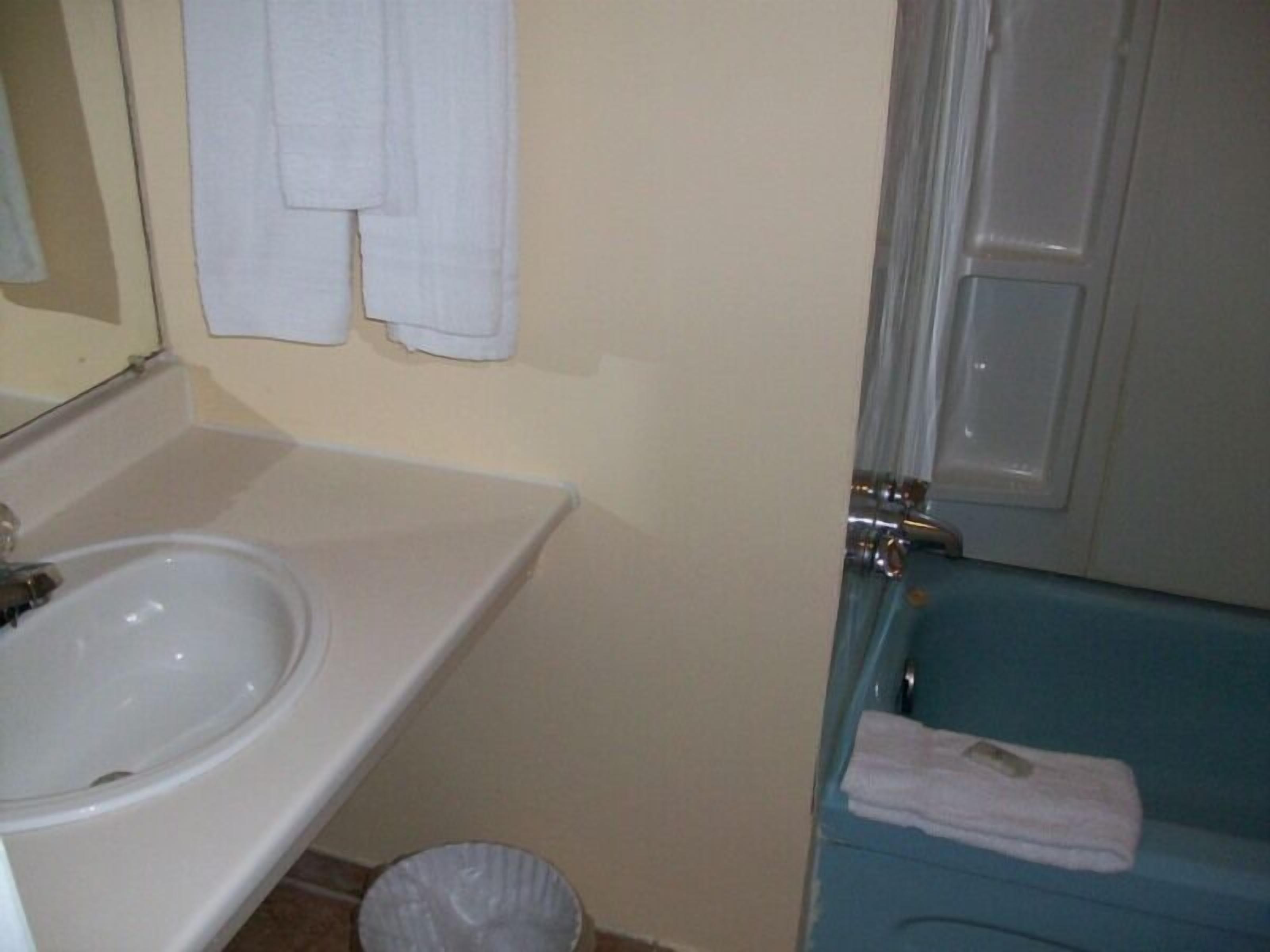 A bathroom with a white sink, a mirror, and a blue bathtub. There are white towels hanging on the wall and a trash can next to the sink.