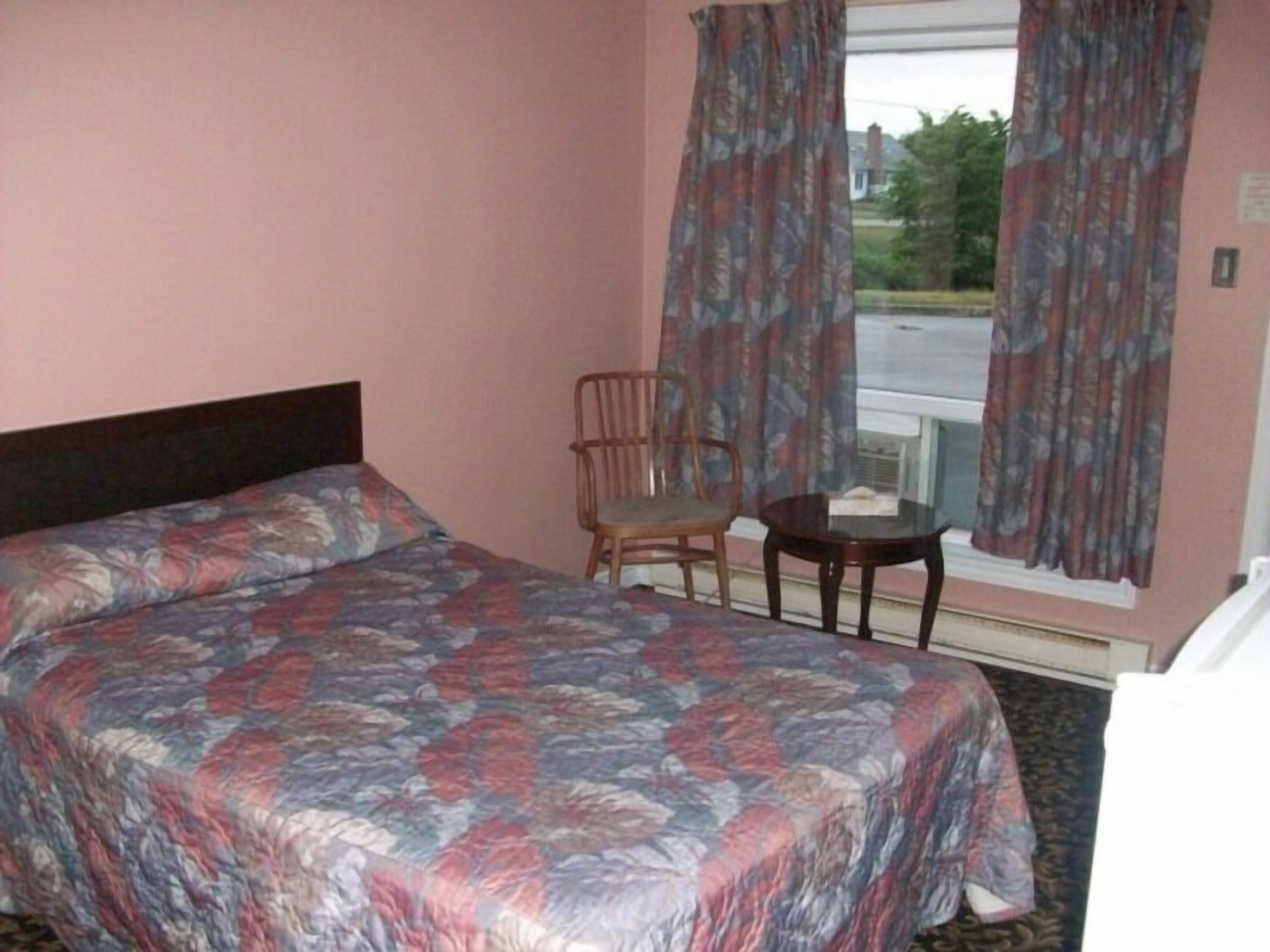 Un dormitorio con cama, una silla, una mesita y una ventana con cortinas.