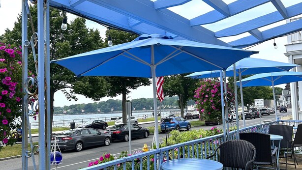 A covered outdoor seating area with blue umbrellas, wicker chairs, and a view of the waterfront. - Harborview Inn (Gloucester)