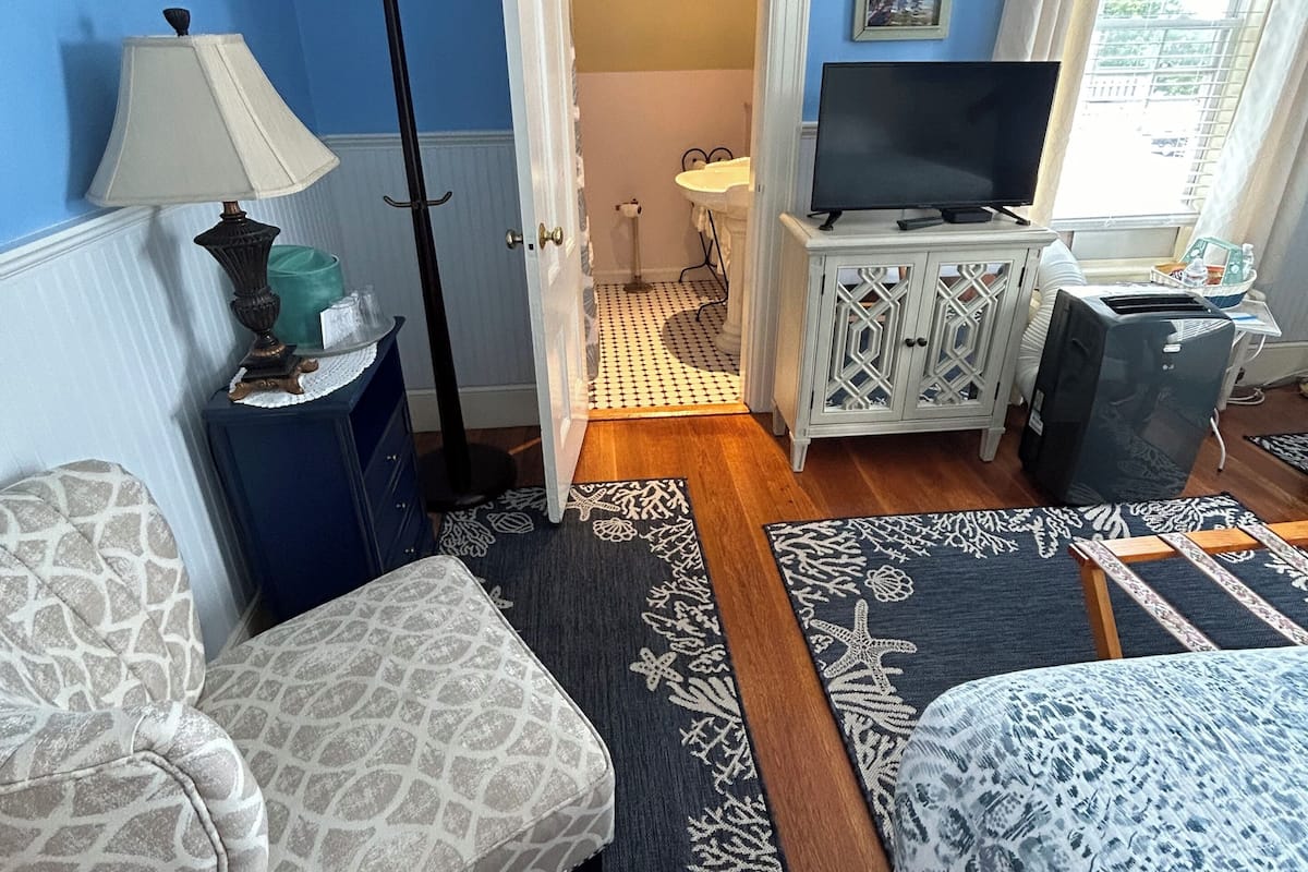 A bedroom with a bed, a chair, a TV, a desk, and a bathroom visible through an open door.
