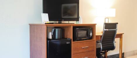 A hotel room with a wooden desk, a black office chair, a flat-screen TV, a microwave, a refrigerator, and a lamp.