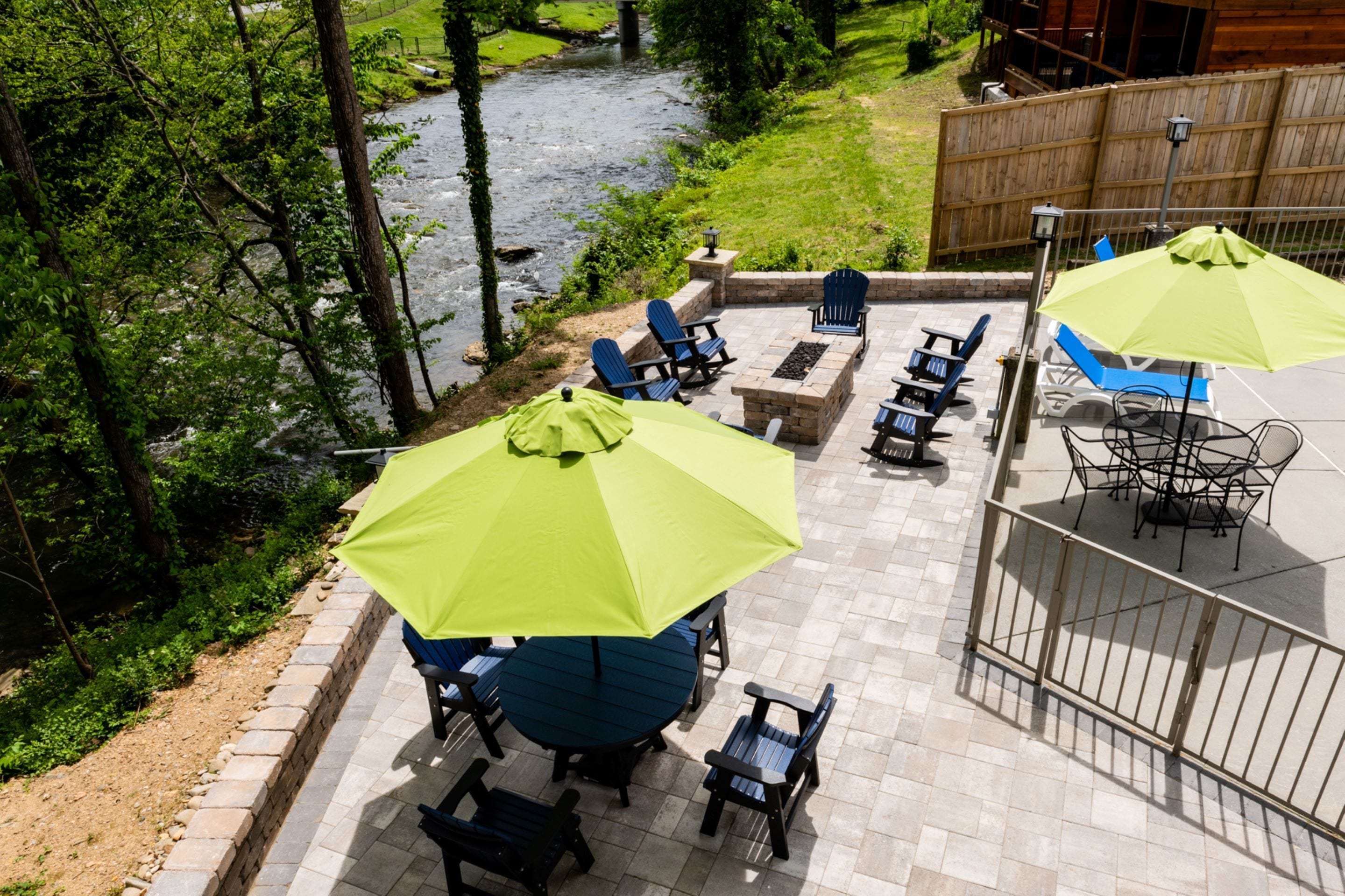 Une terrasse extérieure avec des chaises bleues et des parasols verts, un foyer en pierre et une vue sur une rivière.