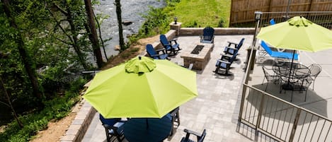 Eine Außenpatio mit blauen Stühlen und grünen Sonnenschirmen, einer Steinkaminfeuerstelle und Blick auf einen Fluss.