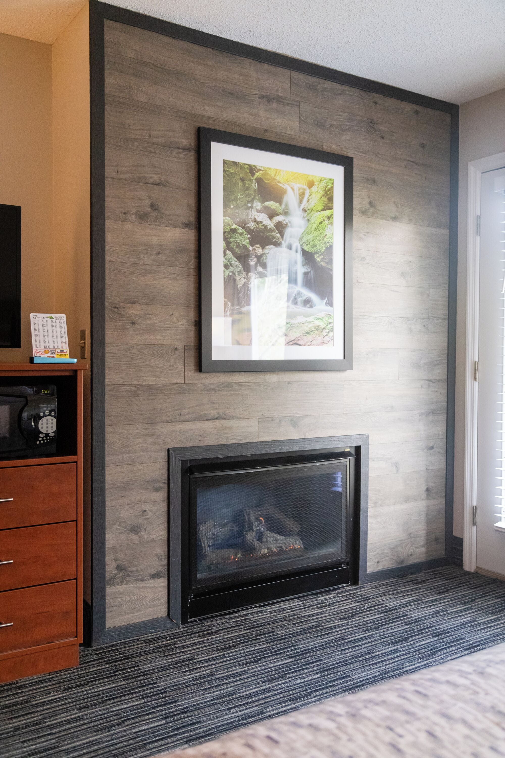 A modern hotel room with a fireplace, a framed picture of a waterfall, and a wooden cabinet with a remote control.