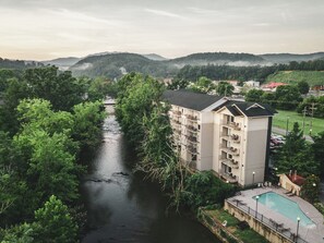 A multi-story hotel with balconies overlooking a river, surrounded by lush greenery and hills.