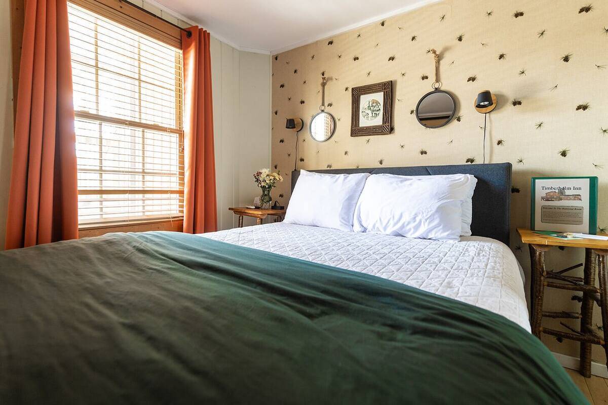 A neatly made bed with a green blanket, a bedside table with a vase of flowers, and a wall with decorative items including a framed picture and two wall-mounted mirrors.