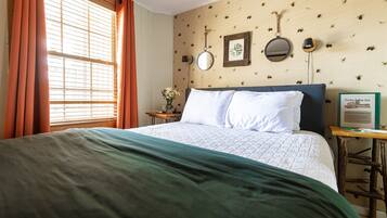 Una cama bien hecha con una manta verde, una mesita de noche con un jarrón de flores, y una pared decorada con objetos como un cuadro enmarcado y dos espejos adosados.