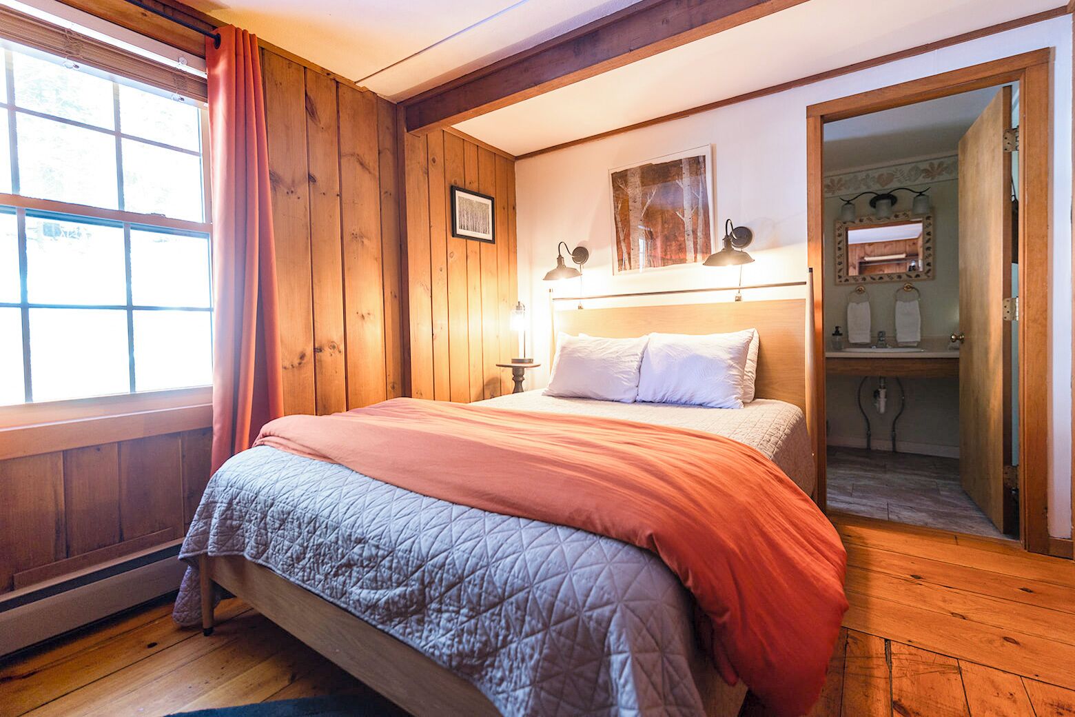 Une chambre à coucher aux murs lambrissés, avec un lit, une table de chevet, un tableau encadré et une salle de bain visible à travers une porte ouverte.