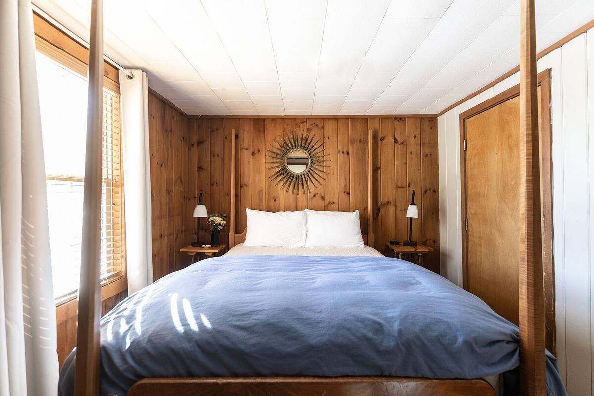 A bedroom with wooden panel walls, a bed with white and blue bedding, two bedside tables with lamps, and a sunburst mirror on the wall.