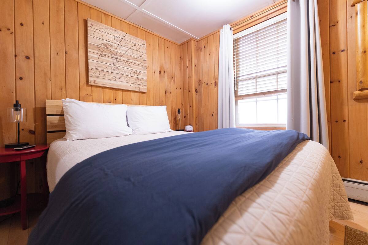 A wooden-paneled room with a bed, a nightstand, a lamp, and a window with blinds.