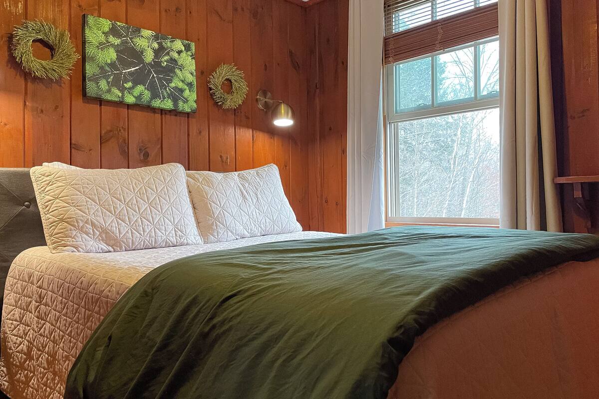 A wooden-paneled bedroom with a bed, two pillows, a green blanket, a wall-mounted lamp, and a window with blinds.