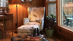 A cozy room with wooden walls, a large window, a plush armchair, a leather ottoman, and a floor lamp. - Timberholm Inn (Stowe)