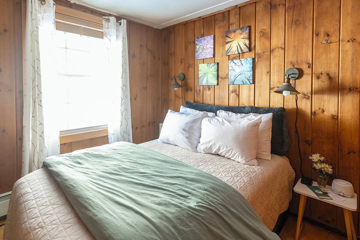 A bedroom with a bed, wooden walls, a window with curtains, and wall-mounted lamps.