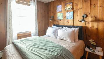 Quarto com cama, paredes de madeira, janela com cortinas e luminárias fixadas na parede.