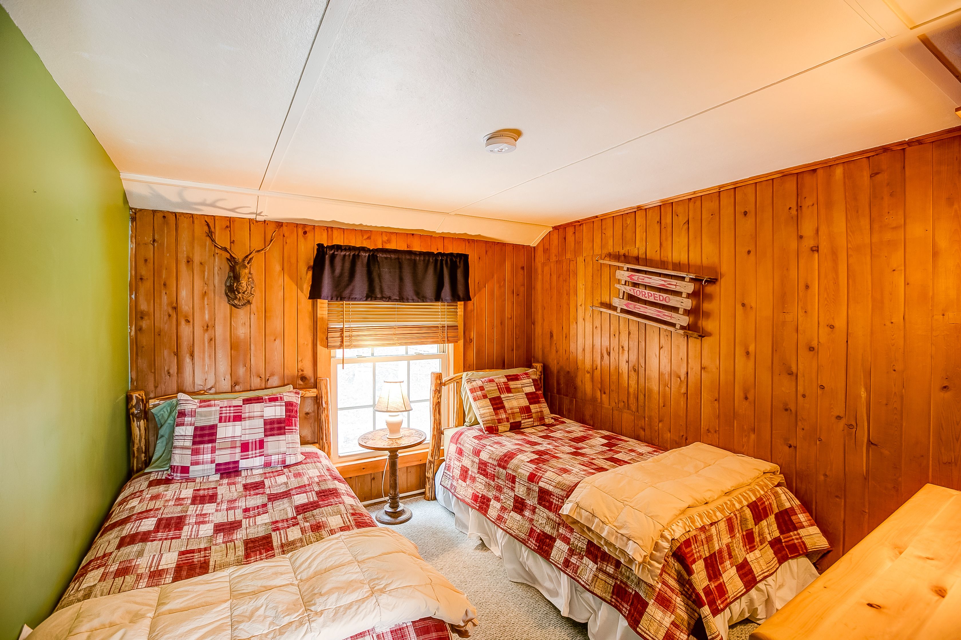 Habitación con dos camas, paredes de madera, una ventana con persianas, una mesita con lámpara y una estantería con libros.
