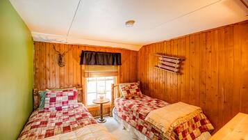 A room with two beds, wooden walls, a window with blinds, a small table with a lamp, and a shelf with books.