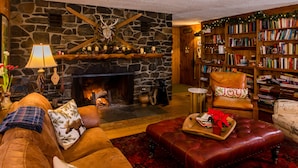 A cozy living room with a stone fireplace, leather furniture, and a bookshelf filled with books. - Timberholm Inn (Stowe)
