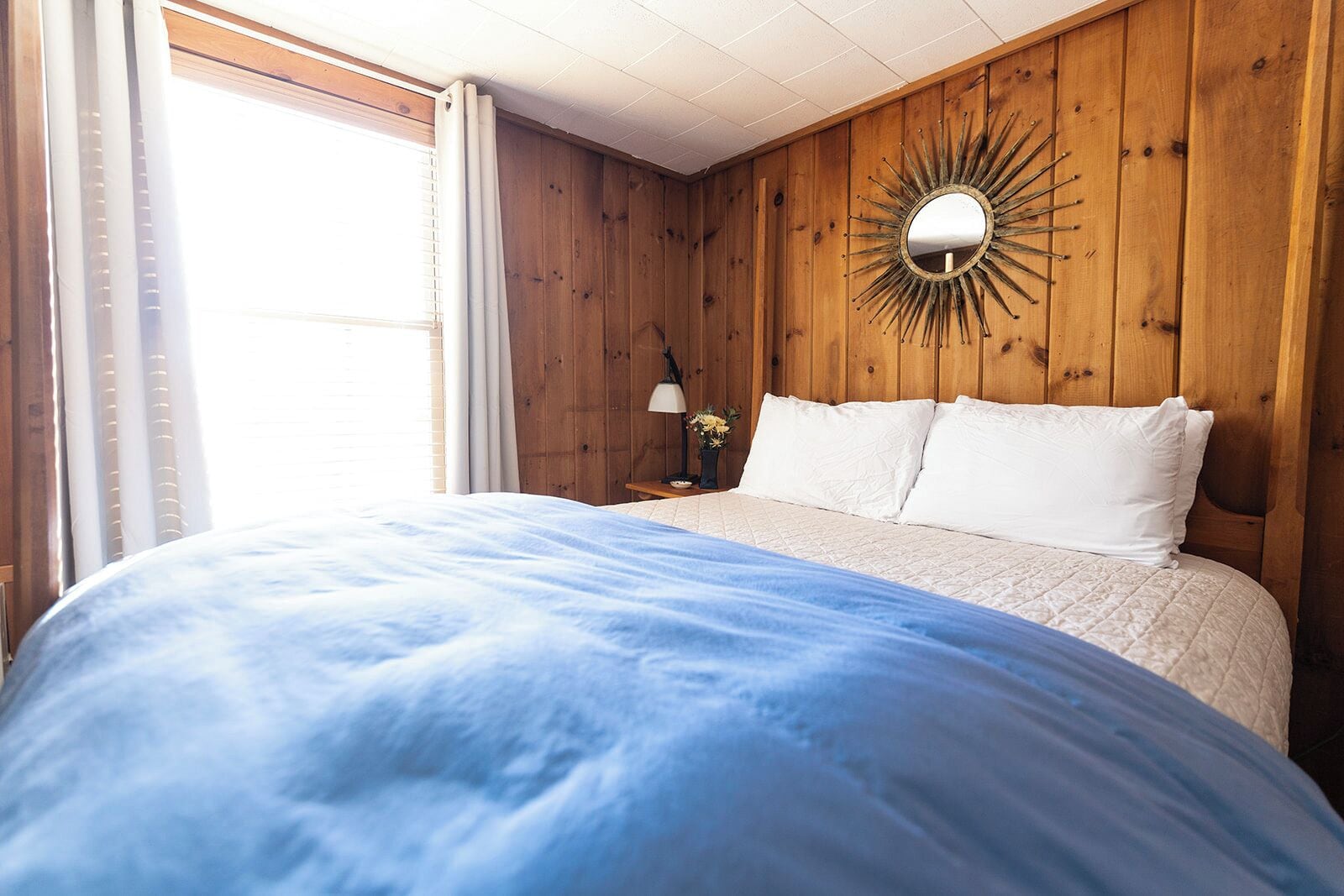 Un dormitori amb un llit gran, parets de fusta, un mirall rodó i una taula petita amb una làmpada.